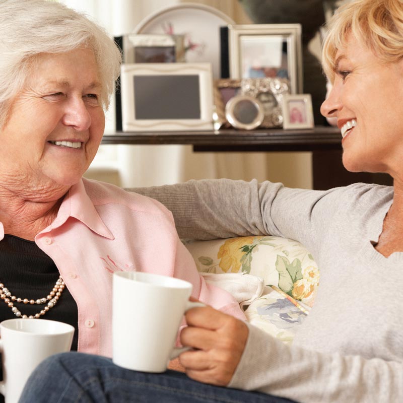 Elderly lady smiling at a younger lady, both holding mugs while sat on sofa