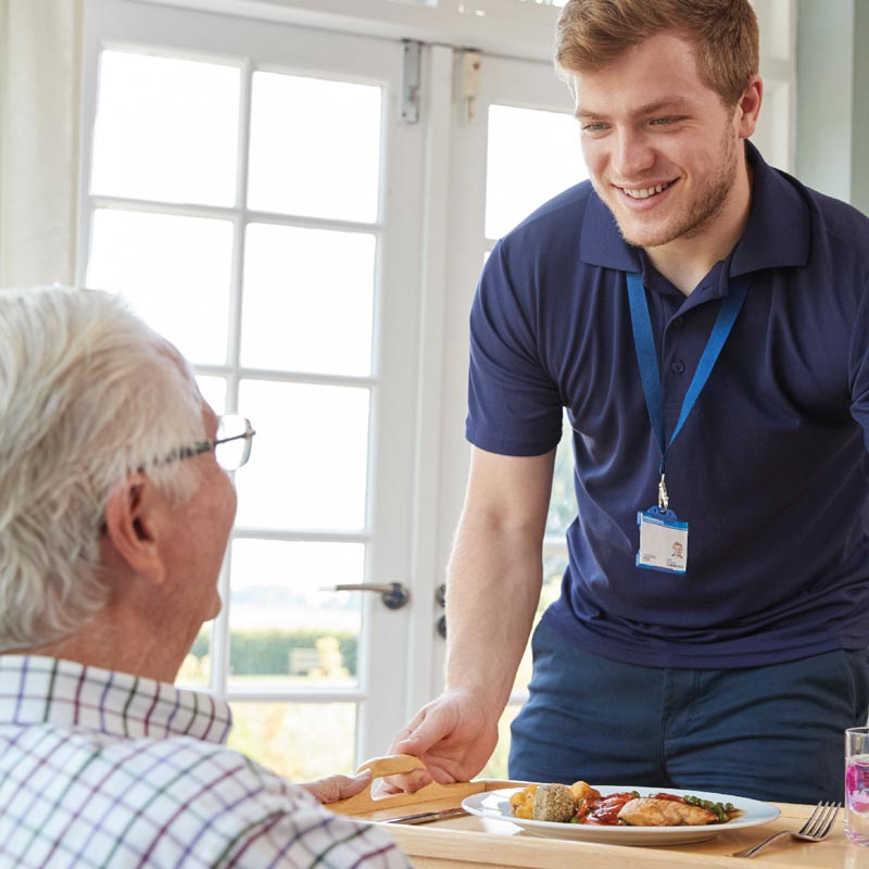 Elderly man sat in foreground, looking up at carer who is smiling