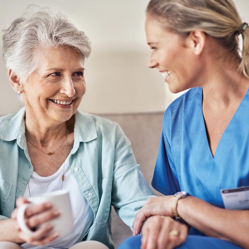 Woman smiling holding a cup of tea, sat with carer who is smiling and holding her hand,.